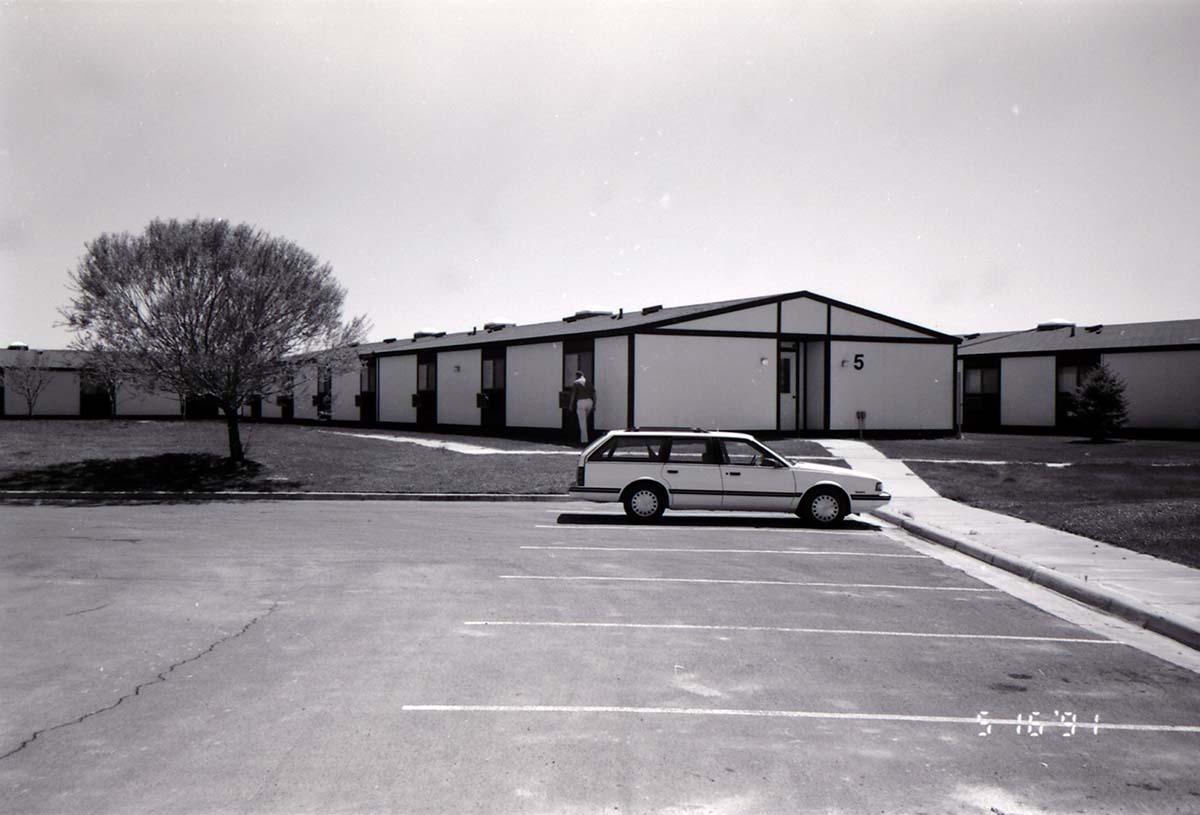 Barracks and parking area with man walking on sidewalk. Building number 5. [Image possibly for comparative housing study]