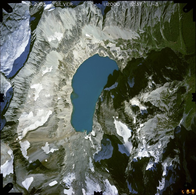 Color aerial photograph of glacier, snowfields, and alpine lake on mountain peak with forested valleys below.