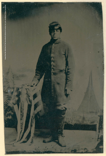 Young man in a full union infantry coat with a kepi on his head. He is wearing high riding boots and his right hand rests on an American flag draped on a table.