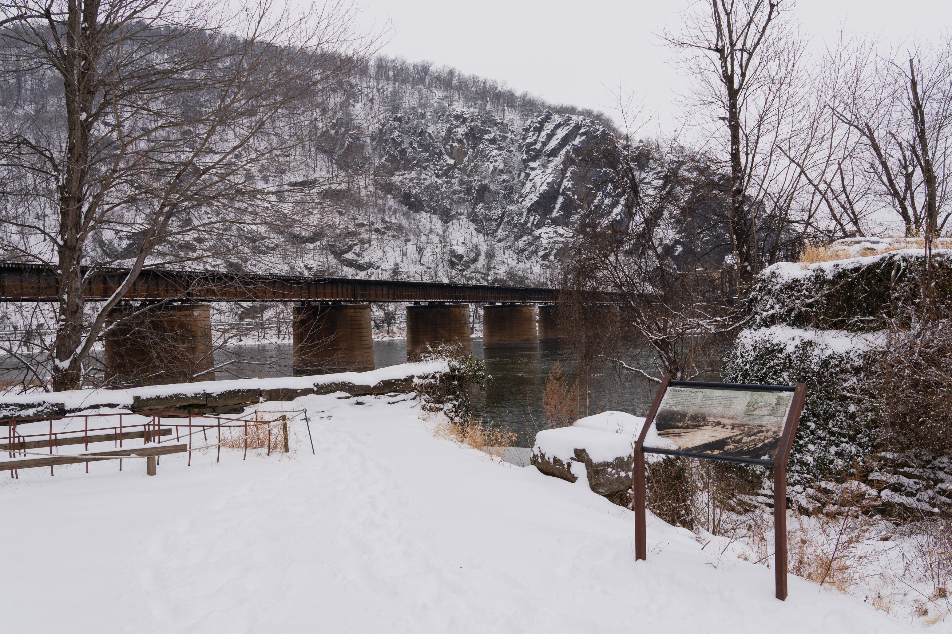 A wayside exhibit sign stands in the foreground, surrounded by snow. In the distance, the B&O Railroad bridge crosses the Potomac River to Maryland Heights, part of the Blue Ridge Mountains.