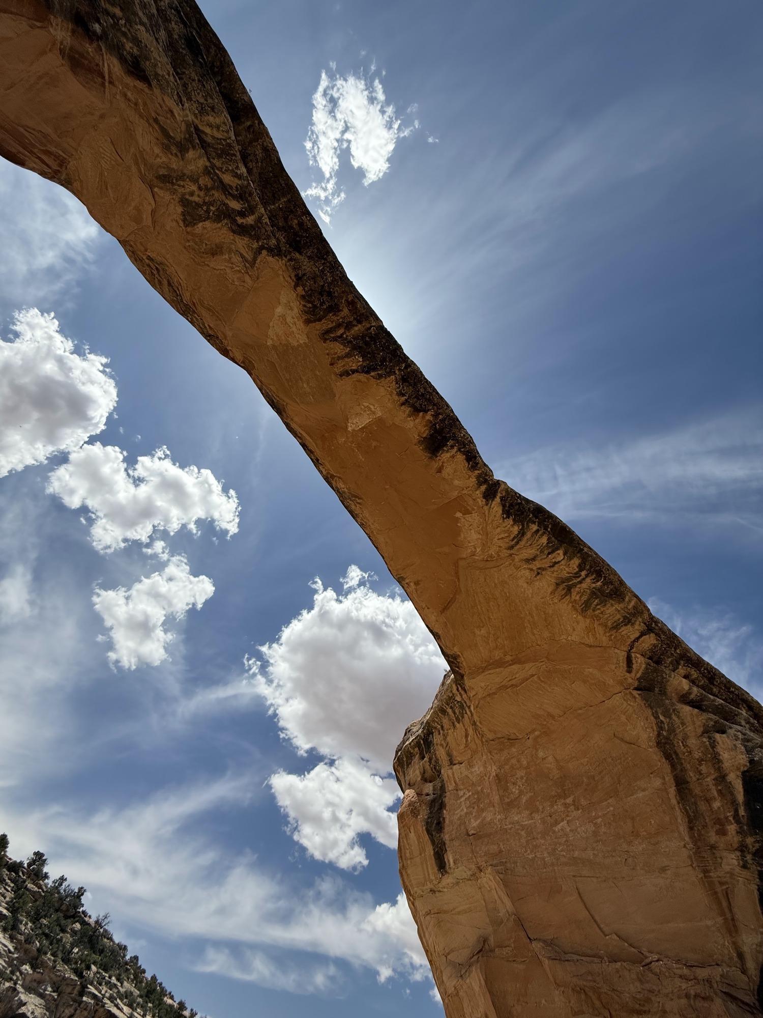 Sun shining out from a natural bridge under blue sky and white puffy clouds.
