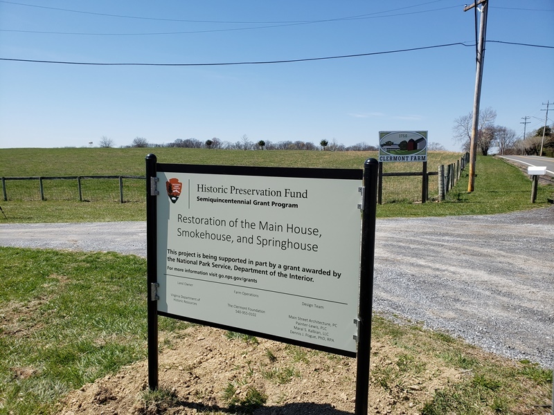 Gravel road with green grass and blue sky on a sunny day. A fenced open field in the background. In the foreground a large sign is visible reading partially Historic Preservation Fund Semiquincentennial Program Restoration of the Main House, Smokehouse, and Springhouse.