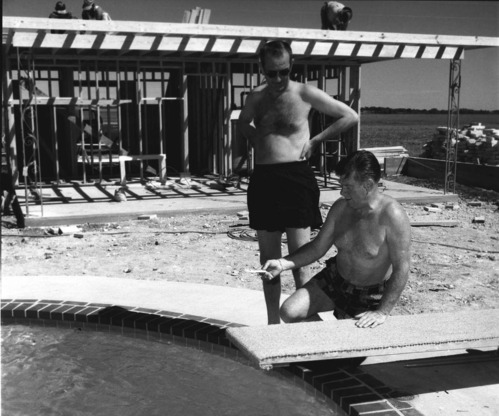 Two men in swim trunks are next to a swimming pool. One is kneeling next to a diving board, holding a thermometer, while the other stands next to him watching. Behind them three men are working on the top of a building frame under construction.
