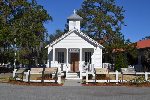 Interpretation signs surround the front porch of the church. A small white, wooden, single-story chapel. The steeple is in the middle of the roof towards the front of the building. Four steps go up to the wooden porch, where a wood front door stands between two windows on either side. 
