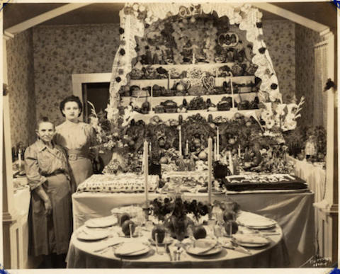 1940: an adult daughter and elderly mother stand beside a ornate and multi-tired altar that stretches from the table to the ceiling. 