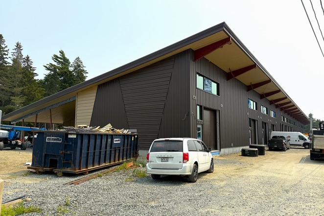 A brown building with a slanted roof under construction. A dumpster sits out front, next to a white van.