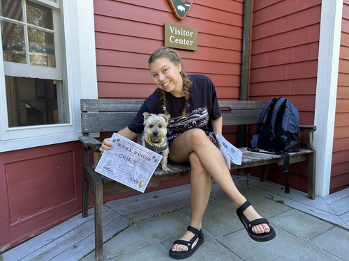 A person holds a certificate and sits on a bench in front of a red building next to a dog