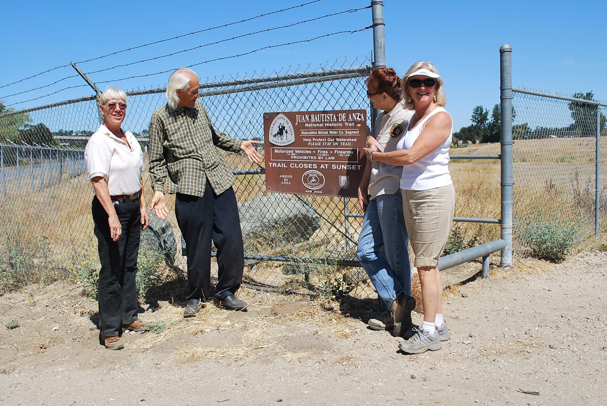 Four people wearing sunglasses stand in front of a cyclone fence with barbed wire that has a Juan Bautista de Anza trail information sign attached