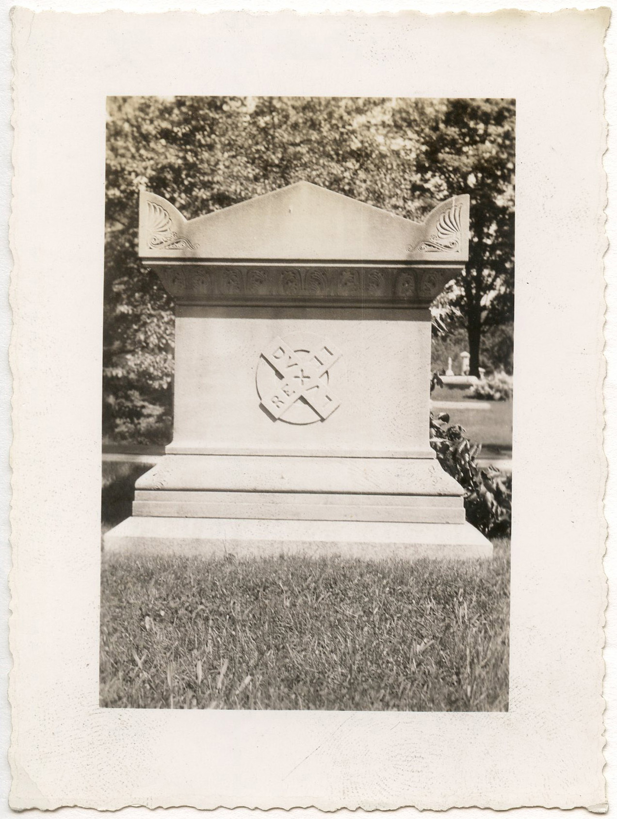 Photograph of Henry Longfellow's grave. View of short side of stone casket-shaped tomb with carved symbol. The symbol is in the shape of an X with a letter X at the center. The letter "X" represents the last letter of the words on each of the four arms of the symbol. The words read "DUX / REX / LUX / LEX." Grass in foreground. Trees in background. A drying wreath is visible on the right side of the image leaning against the tomb.