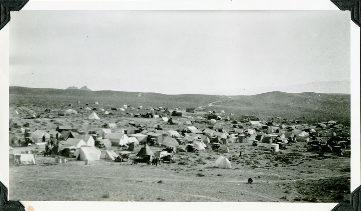 This is an historic black and white photograph from the Scotty's Castle Historic Photograph Collection, Death Valley National Park of encampment of fifty or more mostly tents and some wood shacks. Numerous vehicles, nearly one for every tent or shack. Tents and shacks scattered into the distance. Barren, fairly flat hillside rises into the distance. Double, rugged peak of mountain beyond.