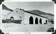 This is an historic black and white photograph from the Scotty's Castle Historic Photograph Collection, Death Valley National Park of Scotty's Castle Stables, looking northeast at north wing. This is immediately after construction with concrete sacks stacking inside and lumber stacking outside. January 14, 1928. Photographed by Mat Roy Thompson.