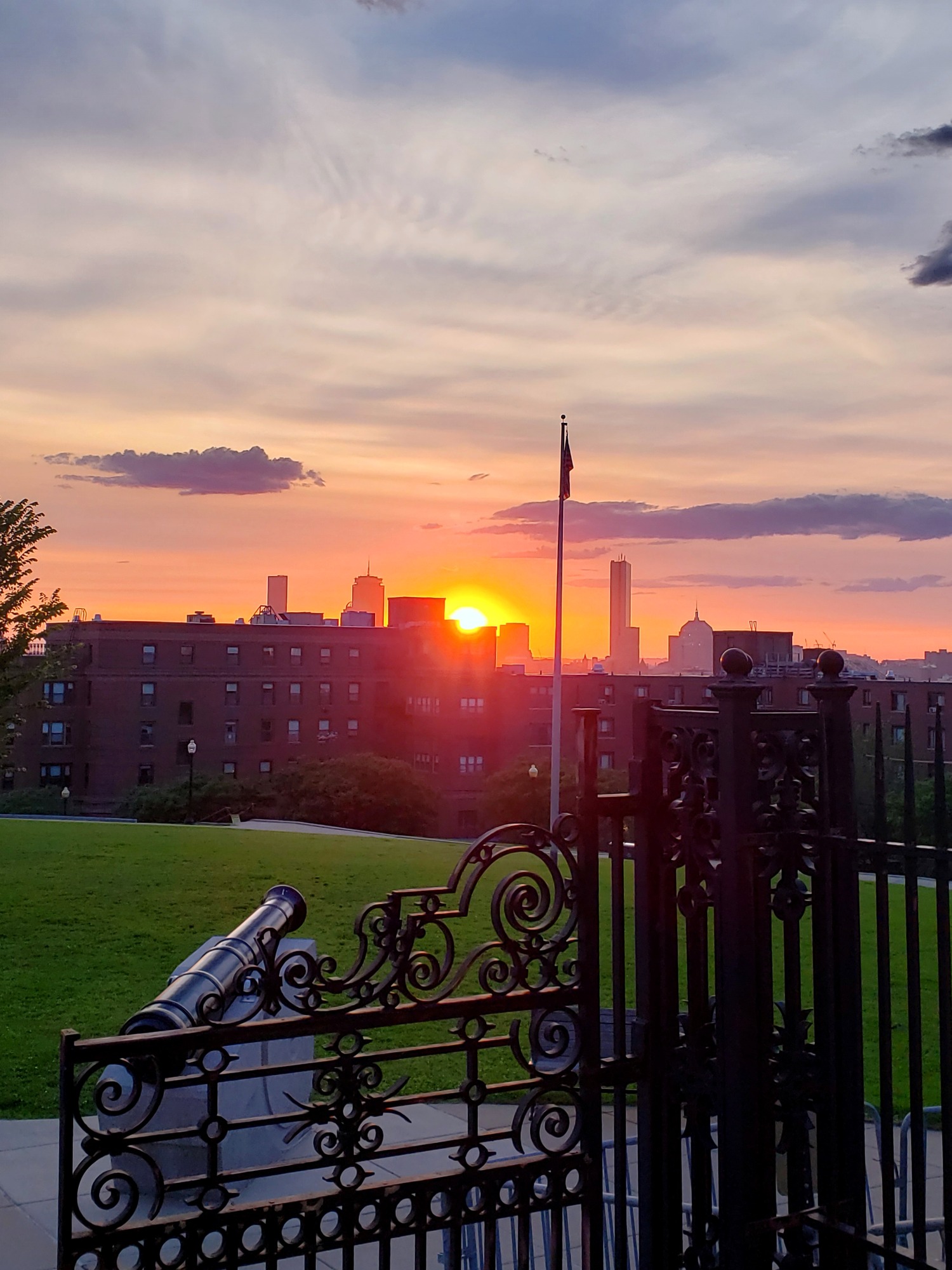sun sets over buildings with Boston skyline in the background. Foreground contains the gates of Dorchester Heights with the cannon and flagpole.