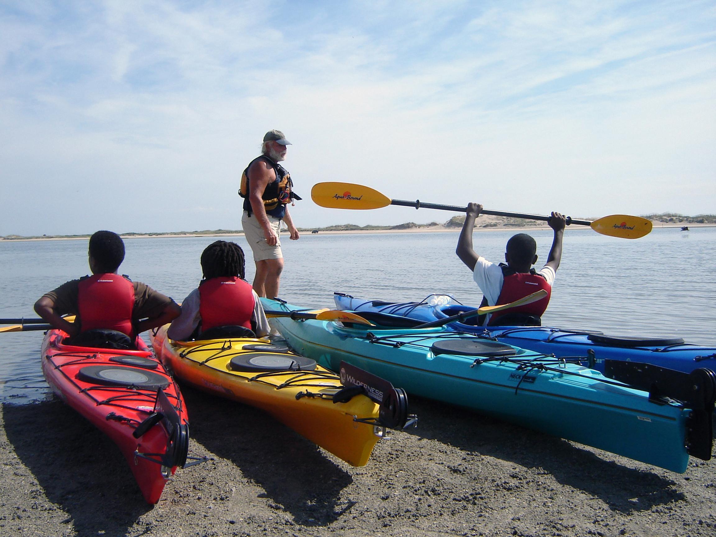 bright kayaks on beach with kids one left is holding paddle in the air while instructor stands in front 