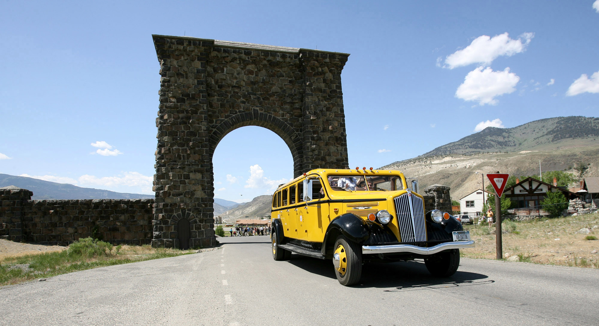 A 1930s style yellow touring bus drives toward camera in front of Roosevelt Arch made out of blocks of basalt rock.