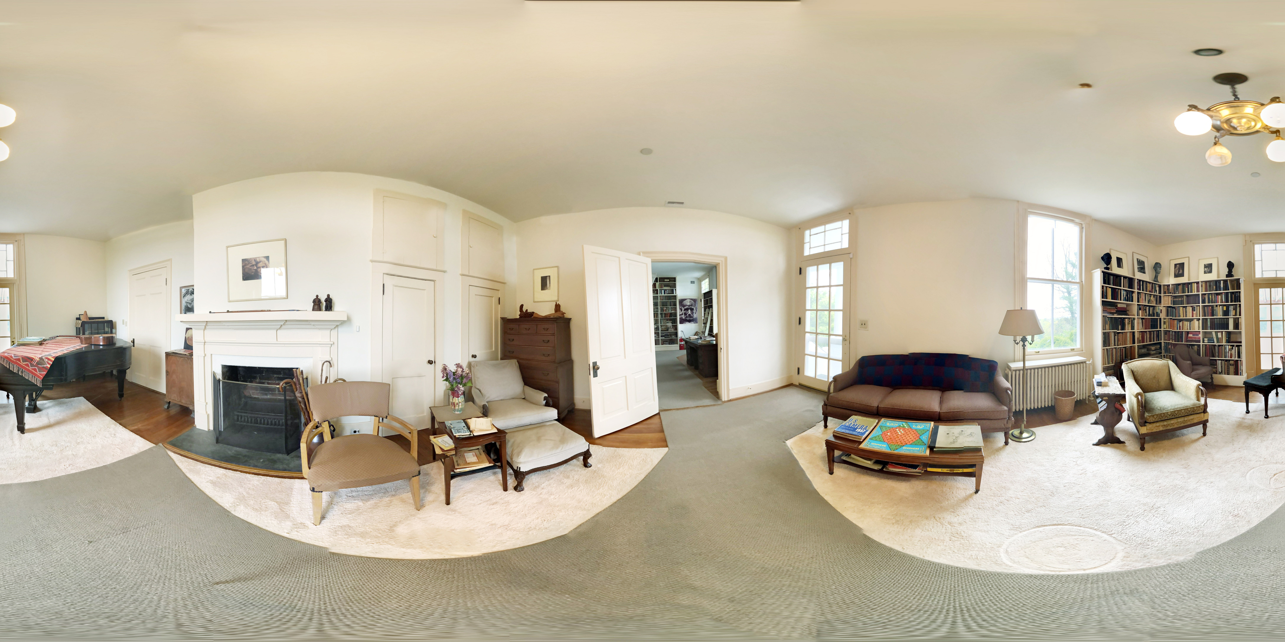 360 photo of the interior of a living room, with a grand piano, sofa and coffee table, chairs, and bookshelves. It is a bright well-lit room with several windows and light painted walls.