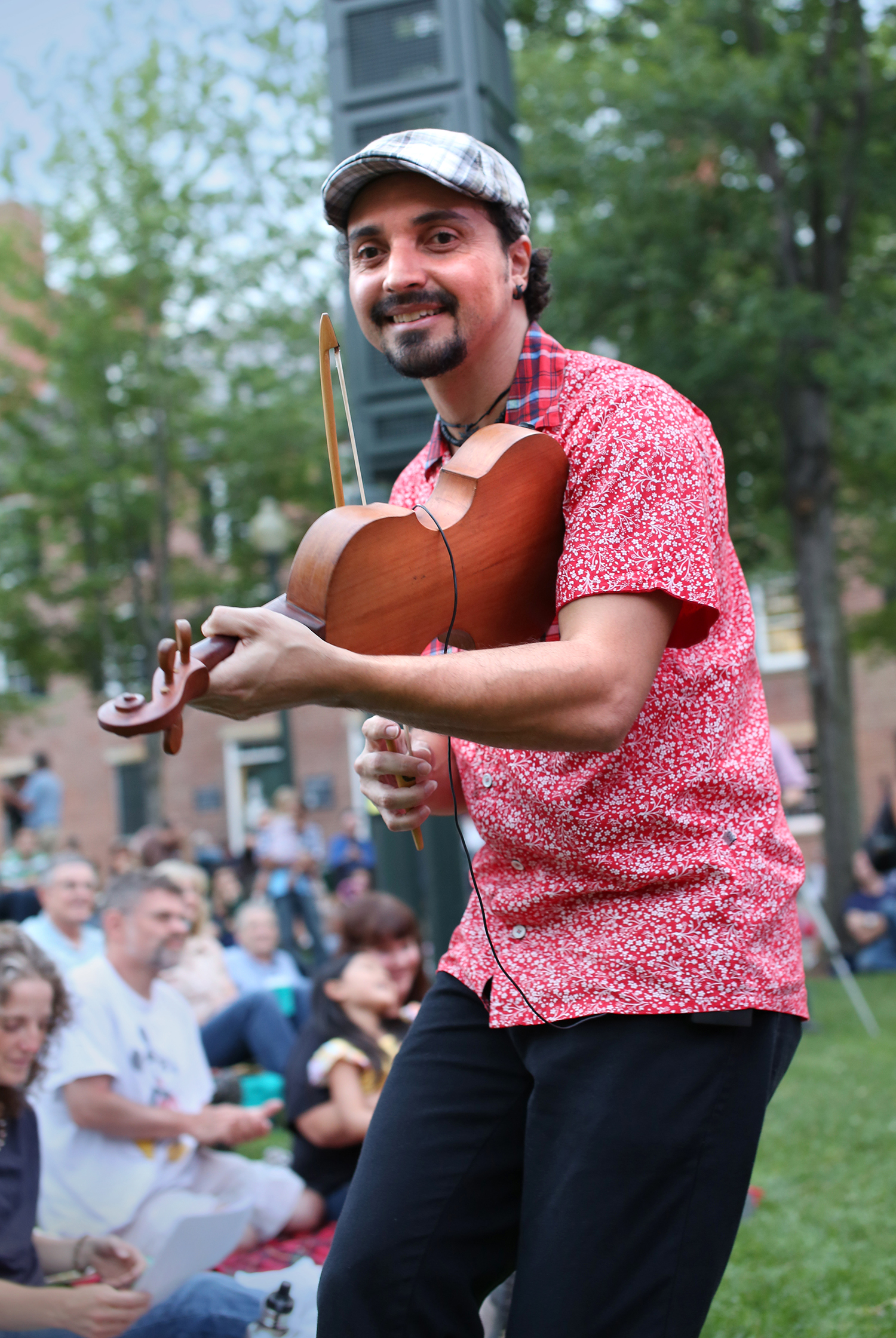 Cláudio Rabeca plays the rabeca, resembling a fiddle, near ground level with the crowd, whom are seated on the grass. Cláudio wears a flat cap, and a shirt of a red paisley pattern and plaid collar. 