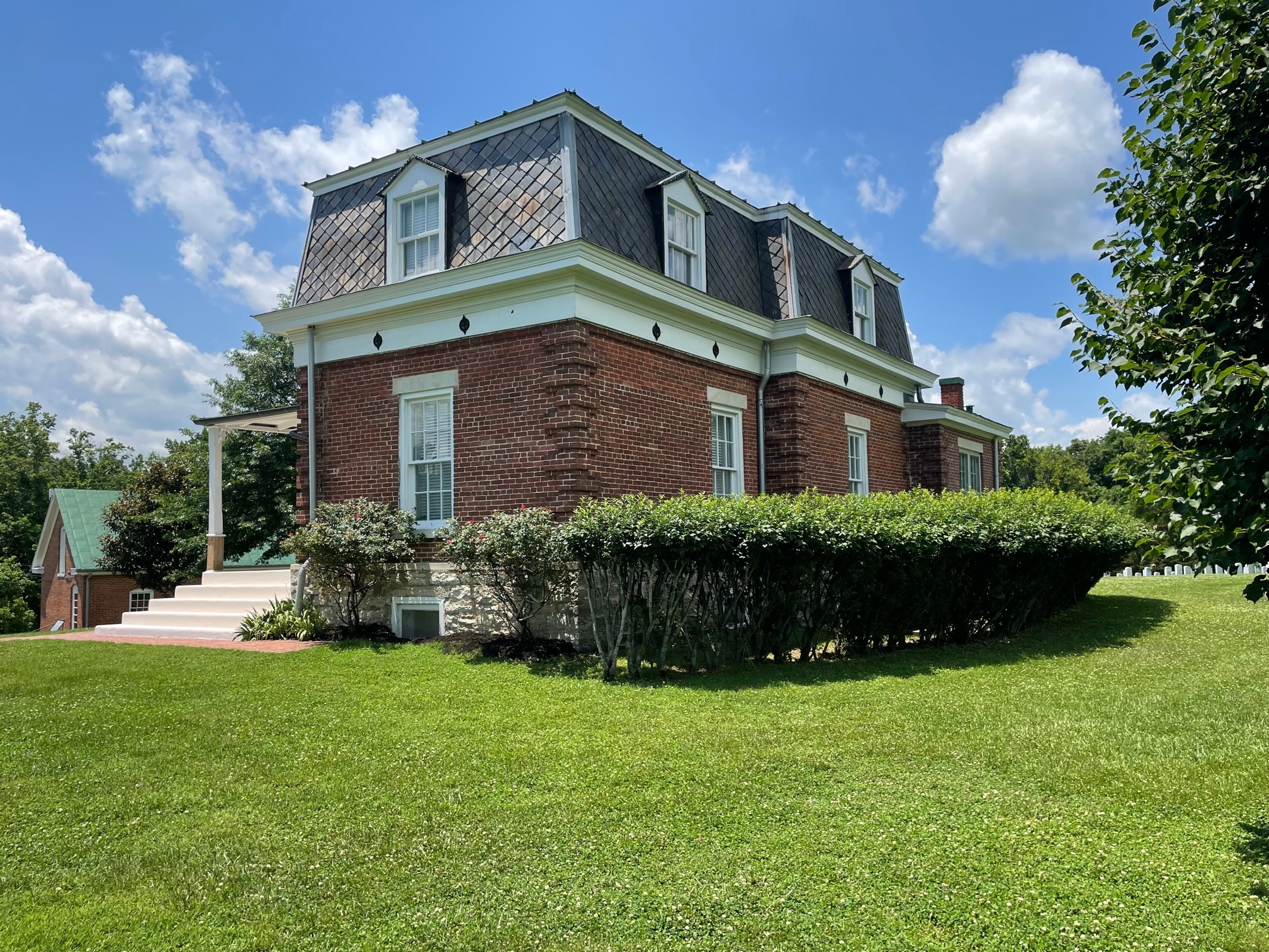 A two-story building has a limestone foundation, brick first story, and mansard roof covered in slate shingles. A hedge grows along the side of the building.  