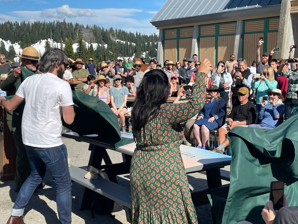 Ada Limon, her brother, and the Superintendent unveil a picnic table in front of a crowd outside in front of a building.