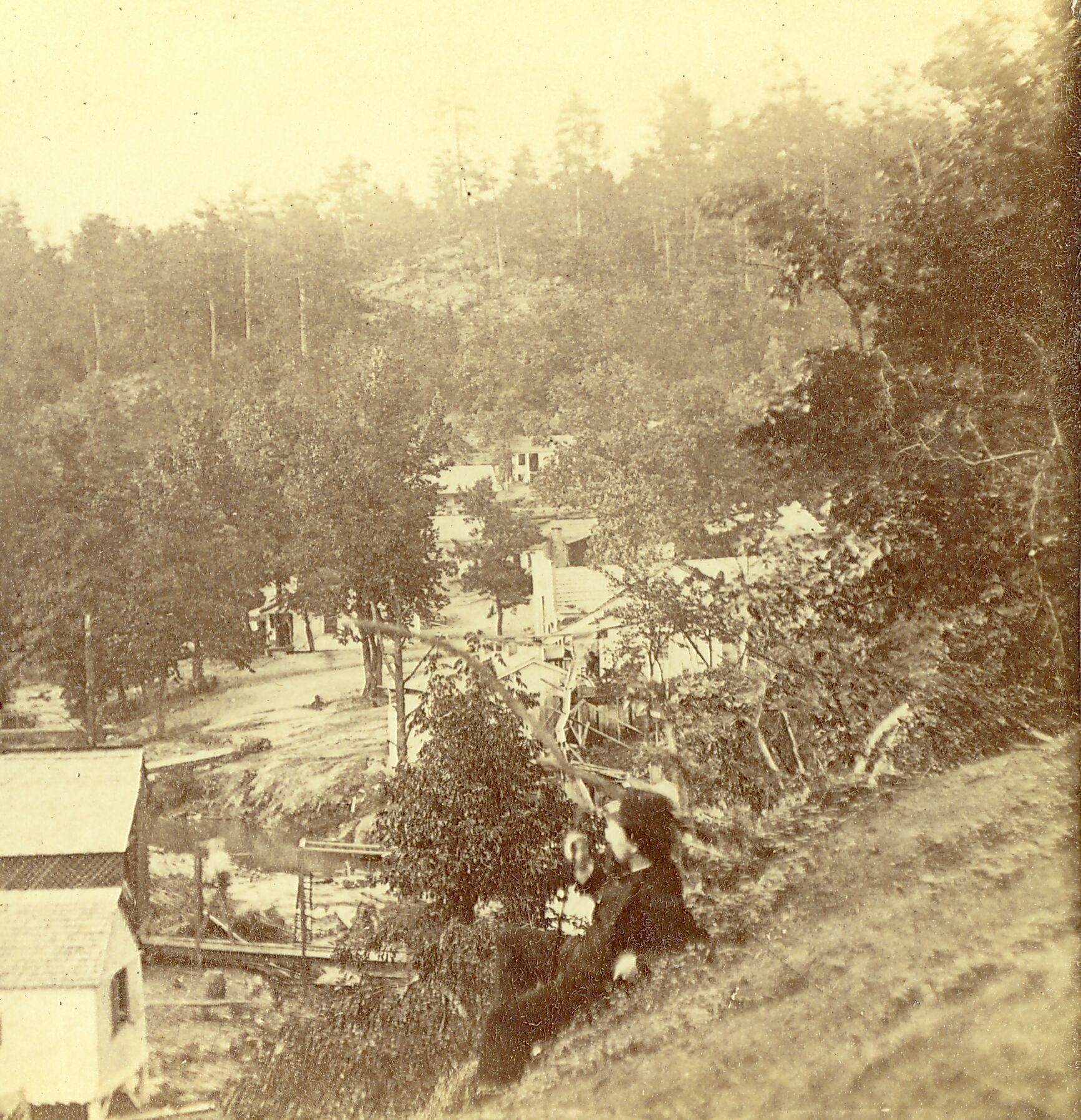 A man sits in the foreground on a hillside overlooking a few small wooden structures and trees. A creek is visible in the background with a plank bridge crossing it.