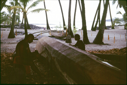 Two men work on carving a waʻa, or Hawaiian canoe, at the beach
