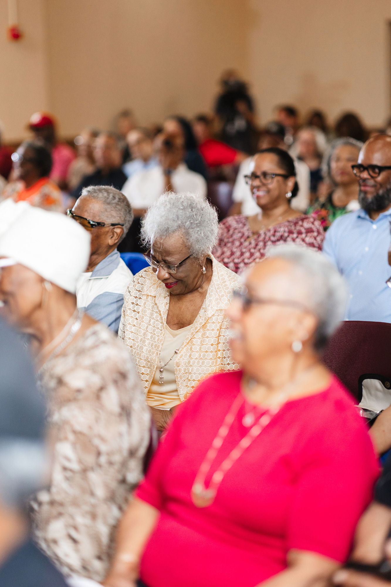 African American adults sit in maroon plastic chairs in a room with hardwood floors the photo focuses on an older african american woman in a light orange jacket chuckling