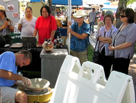 Man throwing a clay bowl at fair as people watch, under a canopy with other booths nearby.