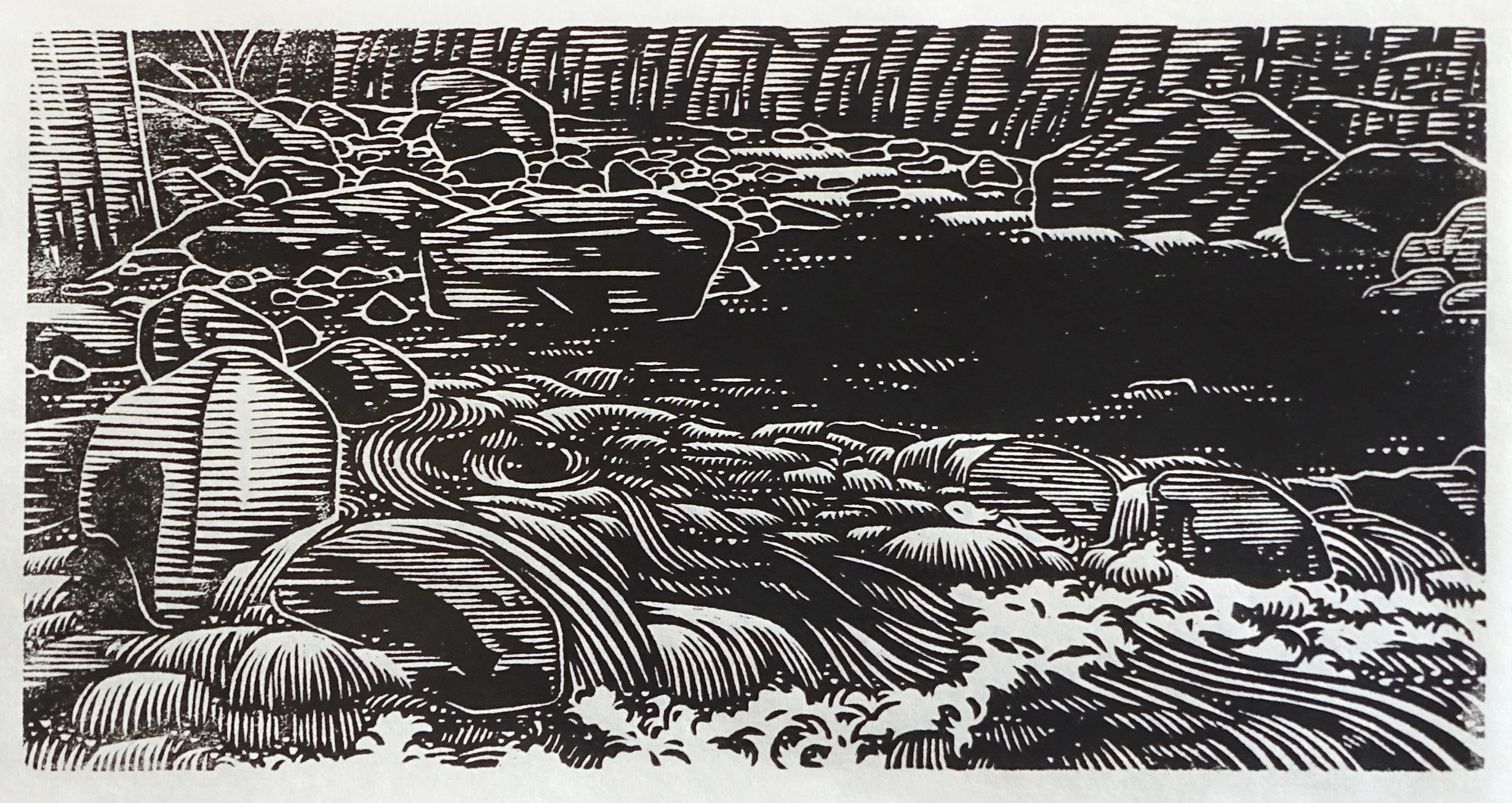 In the foreground, water rushes between boulders, creating white bubbles and rapids. In the middleground, on the right-hand side, a black pool of water sits calmly. In the background, white water is seen flowing over small drops and rapids.