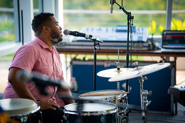 Dominican Republican male speaking into mic, sitting behind the drum set. 