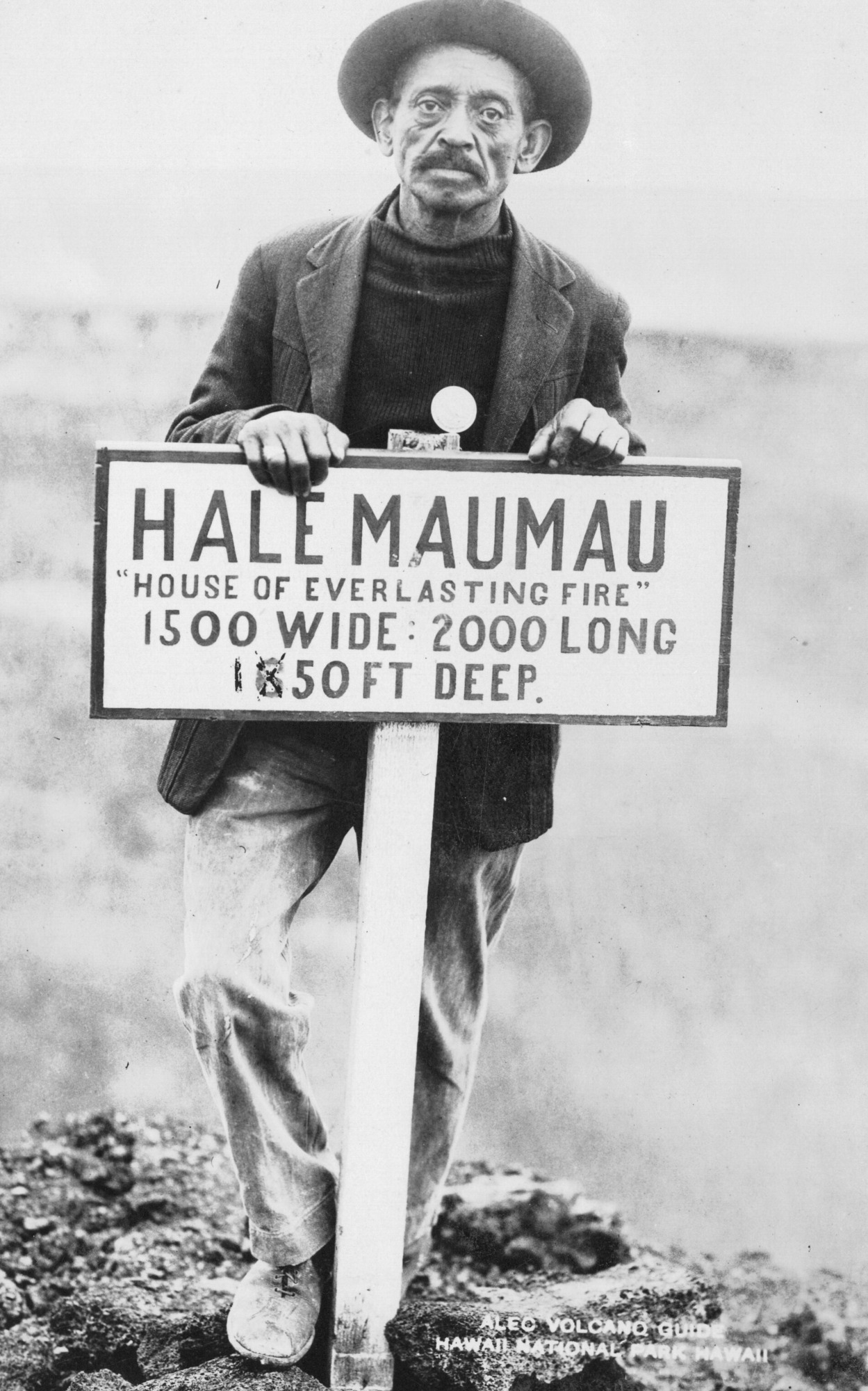 The image is in black and white. Alex, a guide, is posed behind a light-colored sign with dark, all-capitals font that reads, "Halemaumau, "House of Everlasting Fire," 1500 Wide, 2000 Long, 1050 Deep." Alex stands behind the sign, looking at the viewer with his hands resting on top of the sign and his right foot resting higher and his knee bent. He is wearing a sweater, coat, pants, shoes, and a hat. The ground beneath him is lava rock. The background is faint and faded. A caption in the bottom right corner of the image reads, "Alex, Volcano Guide, Hawaii National Park, Hawaii."