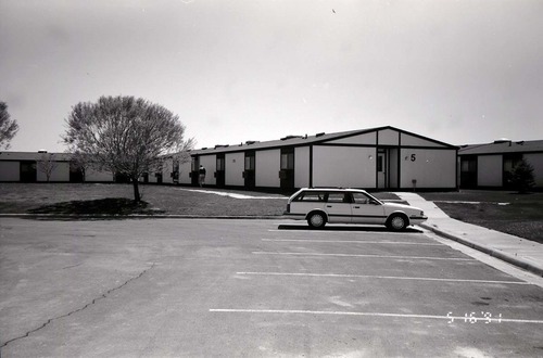 Barracks and parking area with man walking on sidewalk. Building number 5. [Image possibly for comparative housing study]