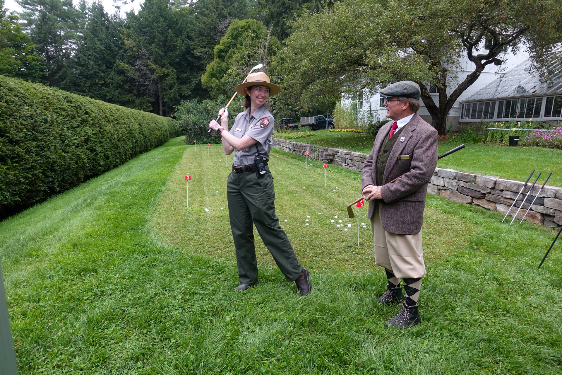 Park Ranger holds golf club and smiles at man dressed in a historic golf outfit in front of a putting green
