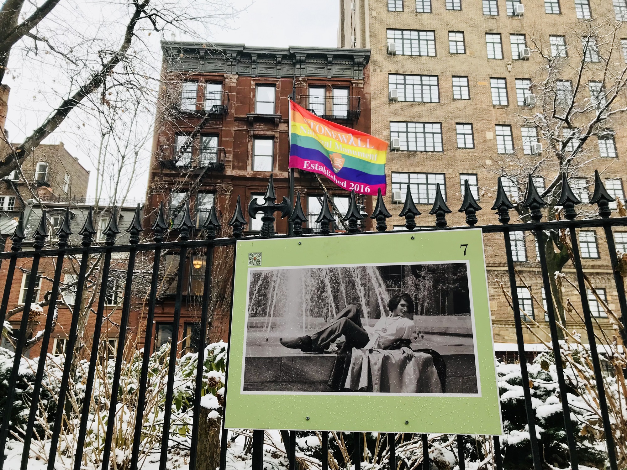 A sign from the fence exhibit at Christopher Park with a photo taken by Kay Tobin of Sylvia Rivera reclining on a fountain in New York City. A rainbow flag proclaiming Stonewall National Monument Established 2016 with the National Park Service logo flies in the background surrounded by city buildings