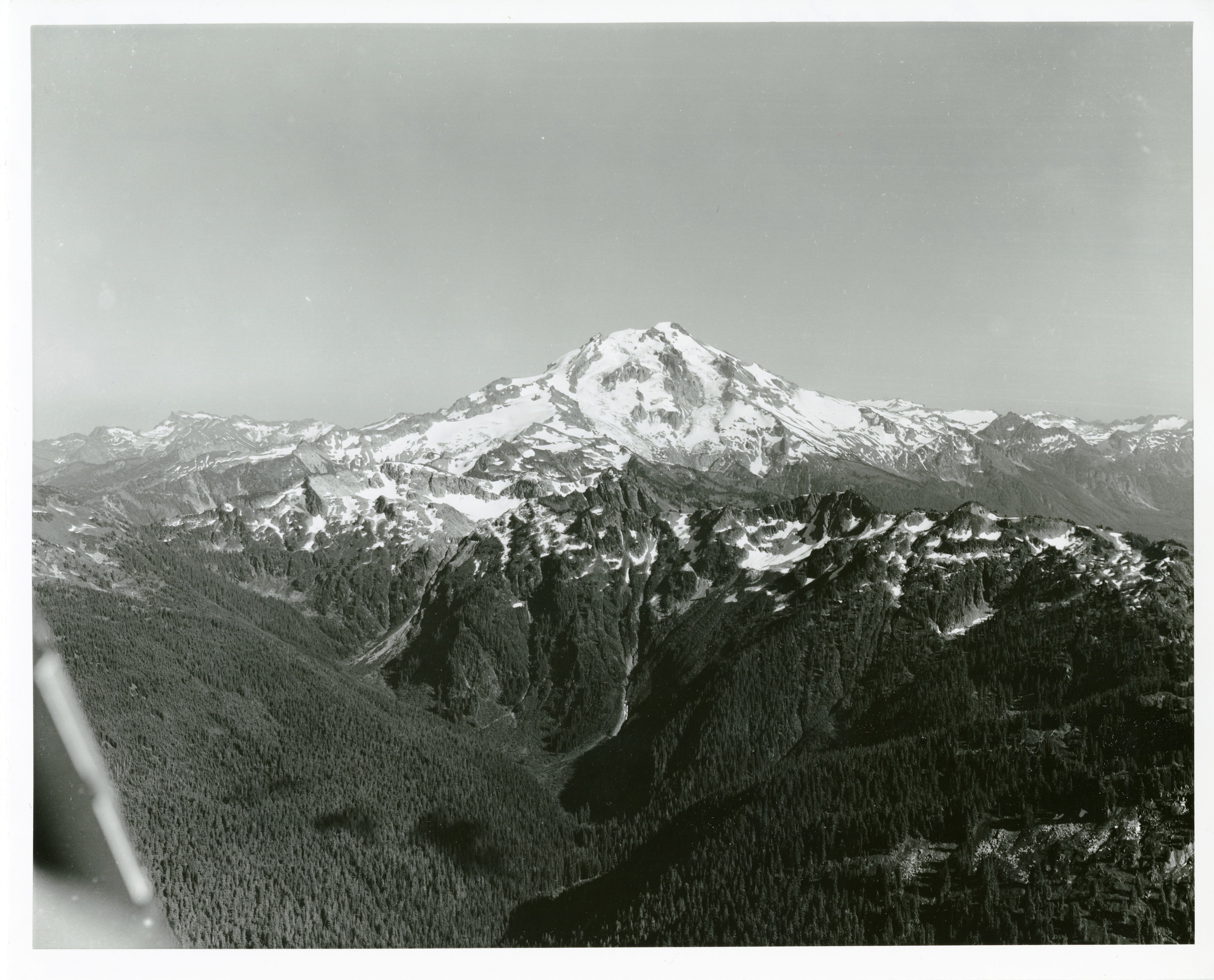 Snow and ice covered peak. There is a smaller, forested ridge closer to the foreground.