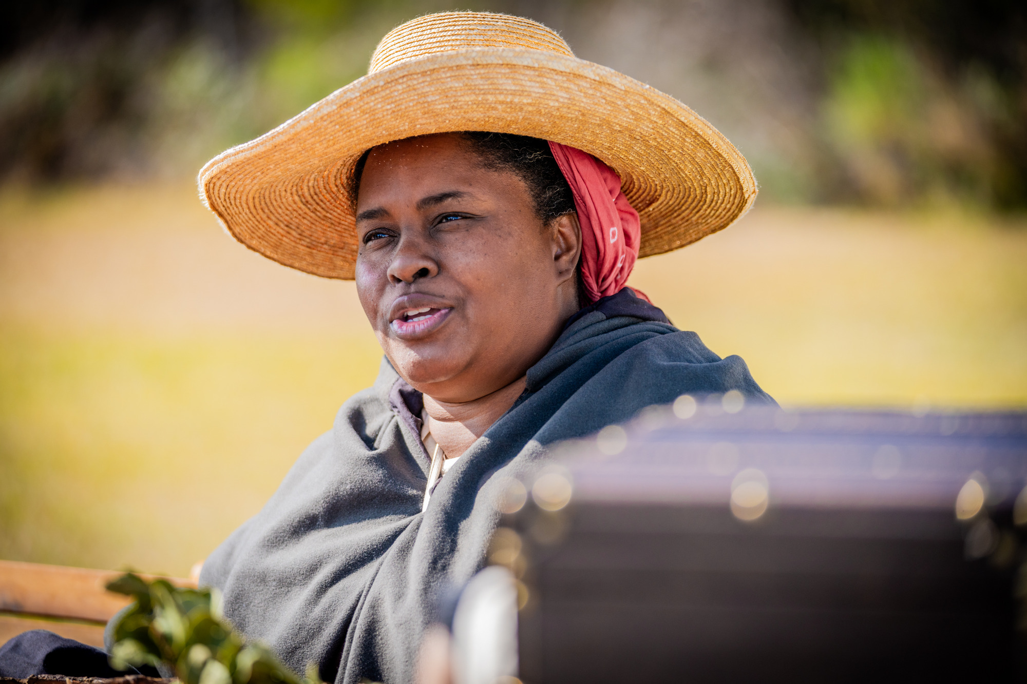 A woman in a straw hat and cloak sits and speaks to someone.