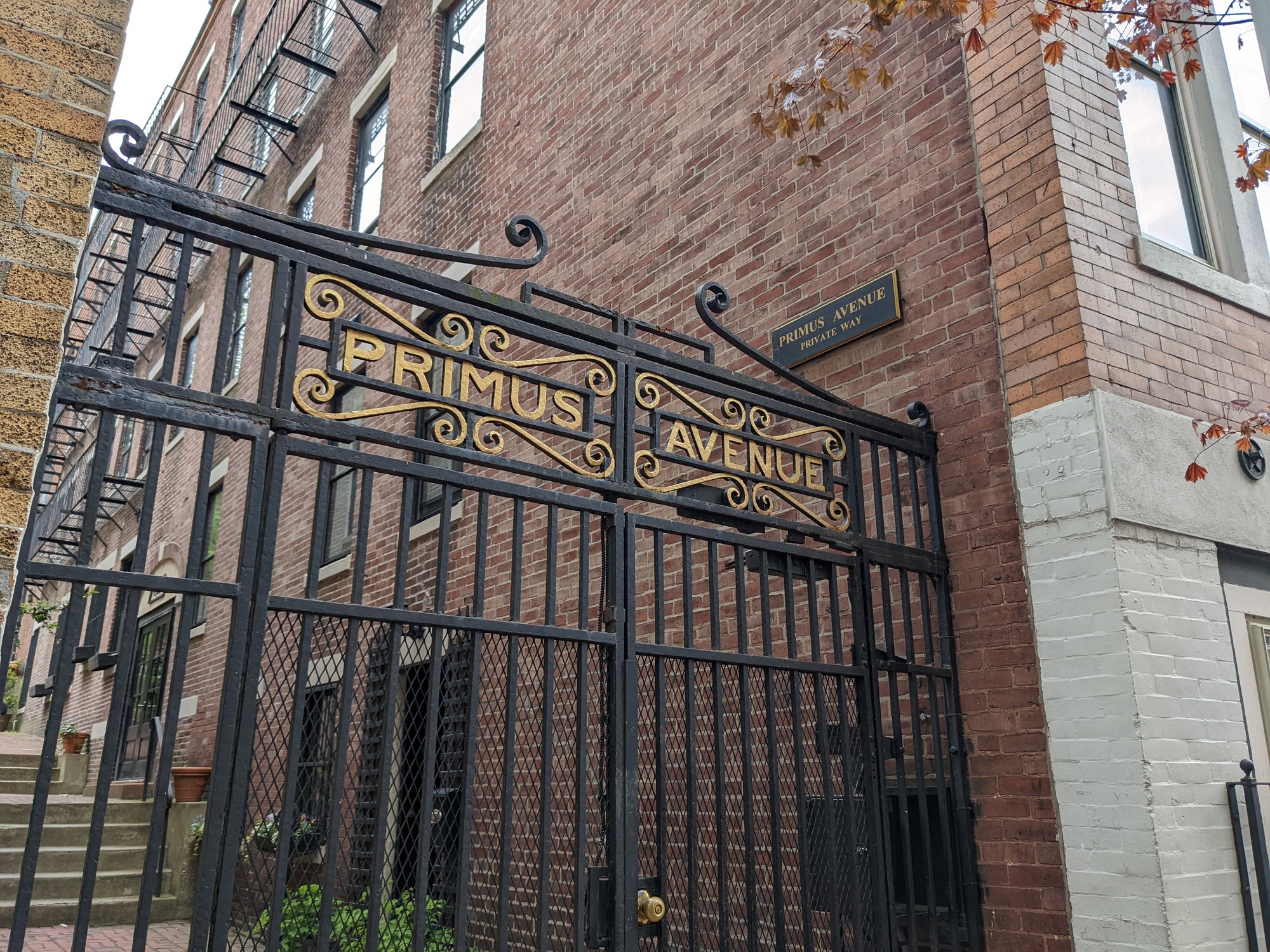 a black iron gate with the words primus avenue sits at the entrance of an alley, a plaque reading Primus Avenue is on a brick wall