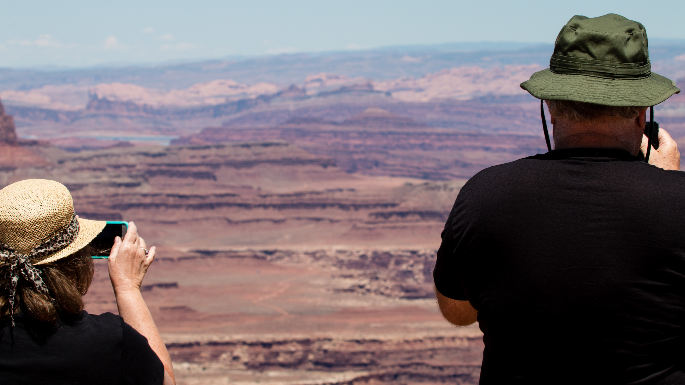 Two visitors take in the view of large canyons in the desert. One visitor is taking photos with their smartphone. 