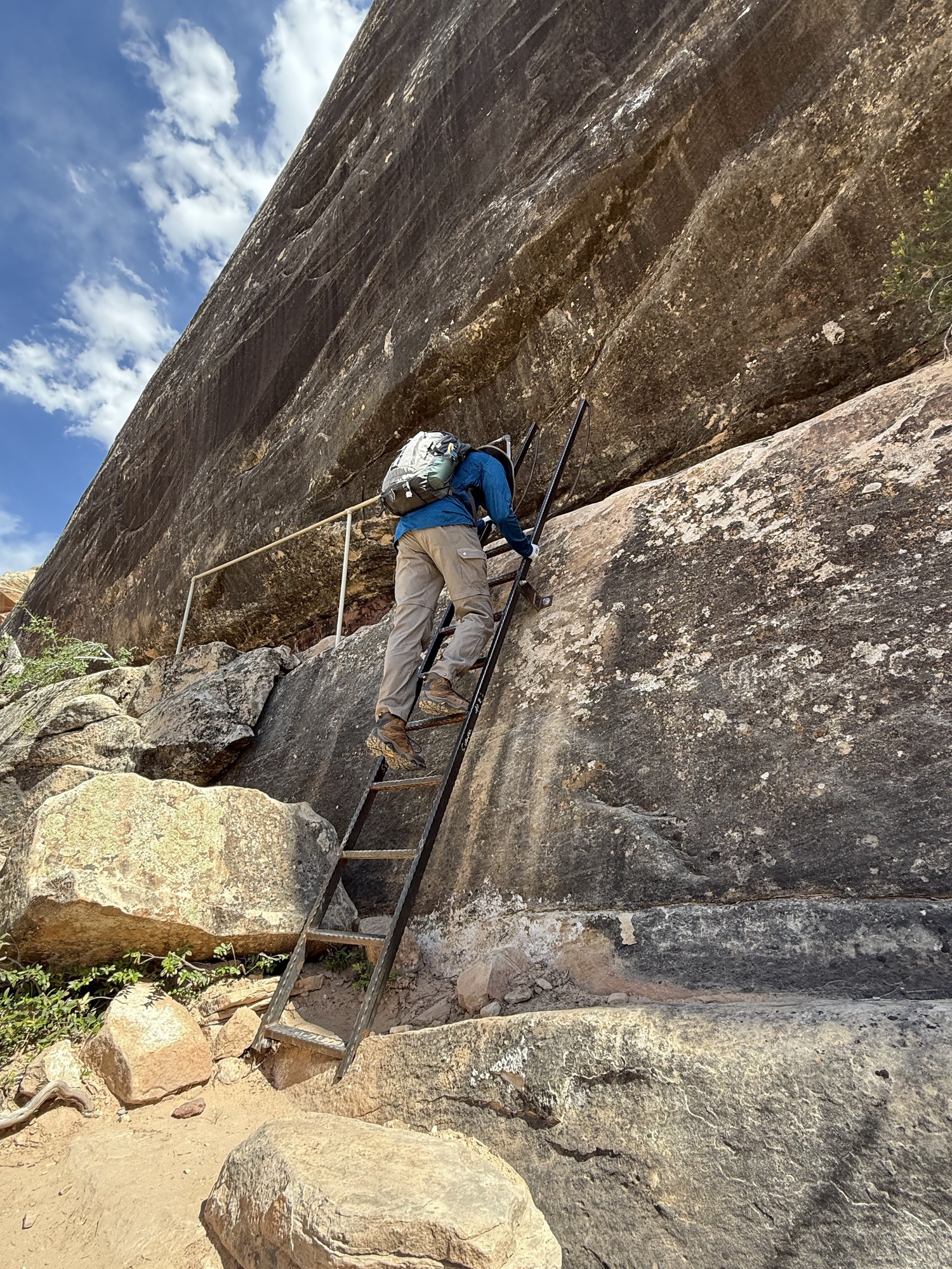 A person climbing down a ladder in a canyon with blue sky and white clouds overhead.