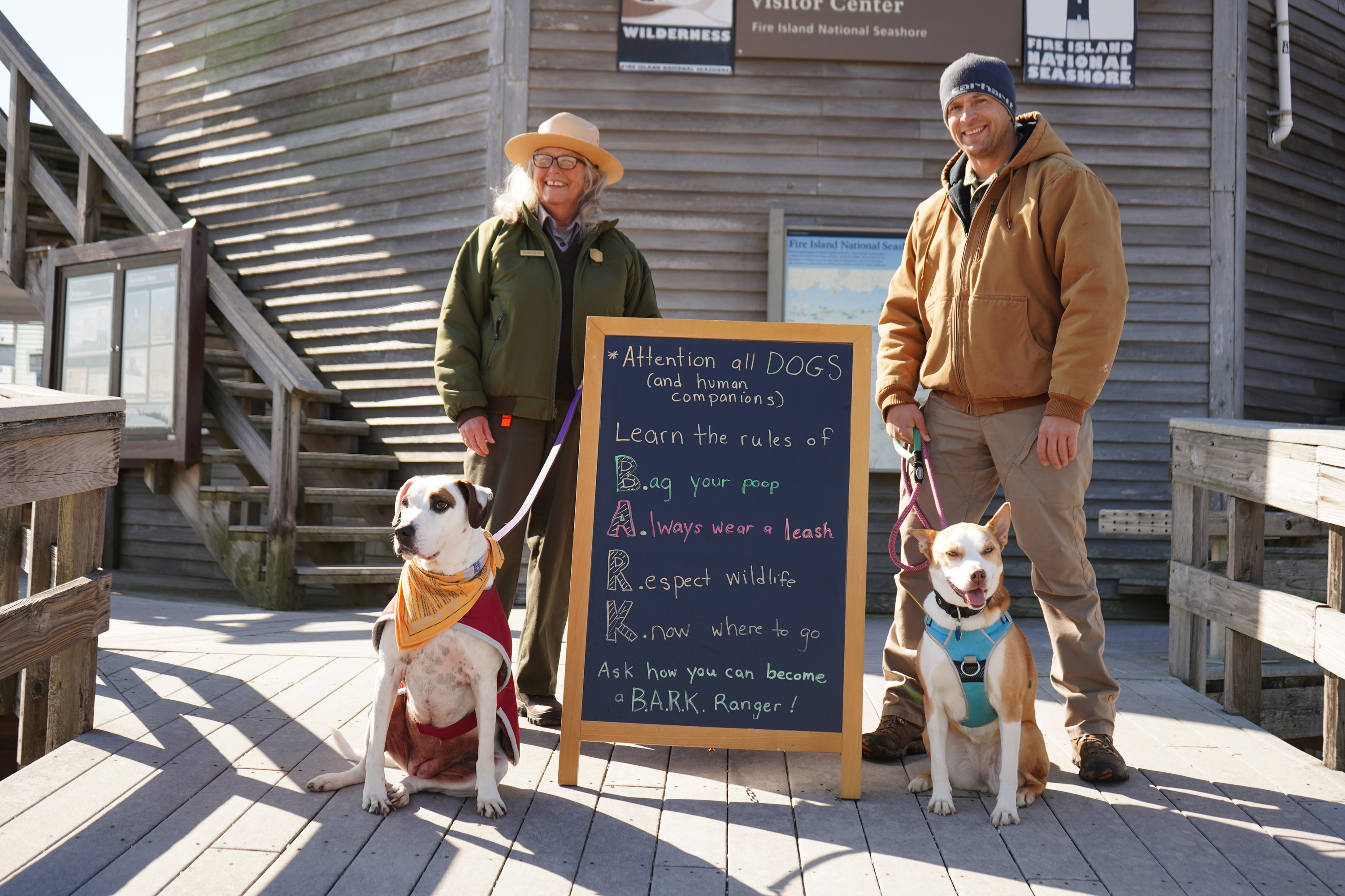 A park ranger and a visitor, each with a leashed dog, pose on a boardwalk near a wooden building.