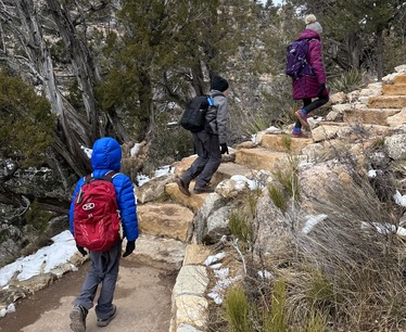 Three Jr. Rangers climb up the many limestone steps of the Island Trail at Walnut Canyon surrounded by snow and juniper trees. 