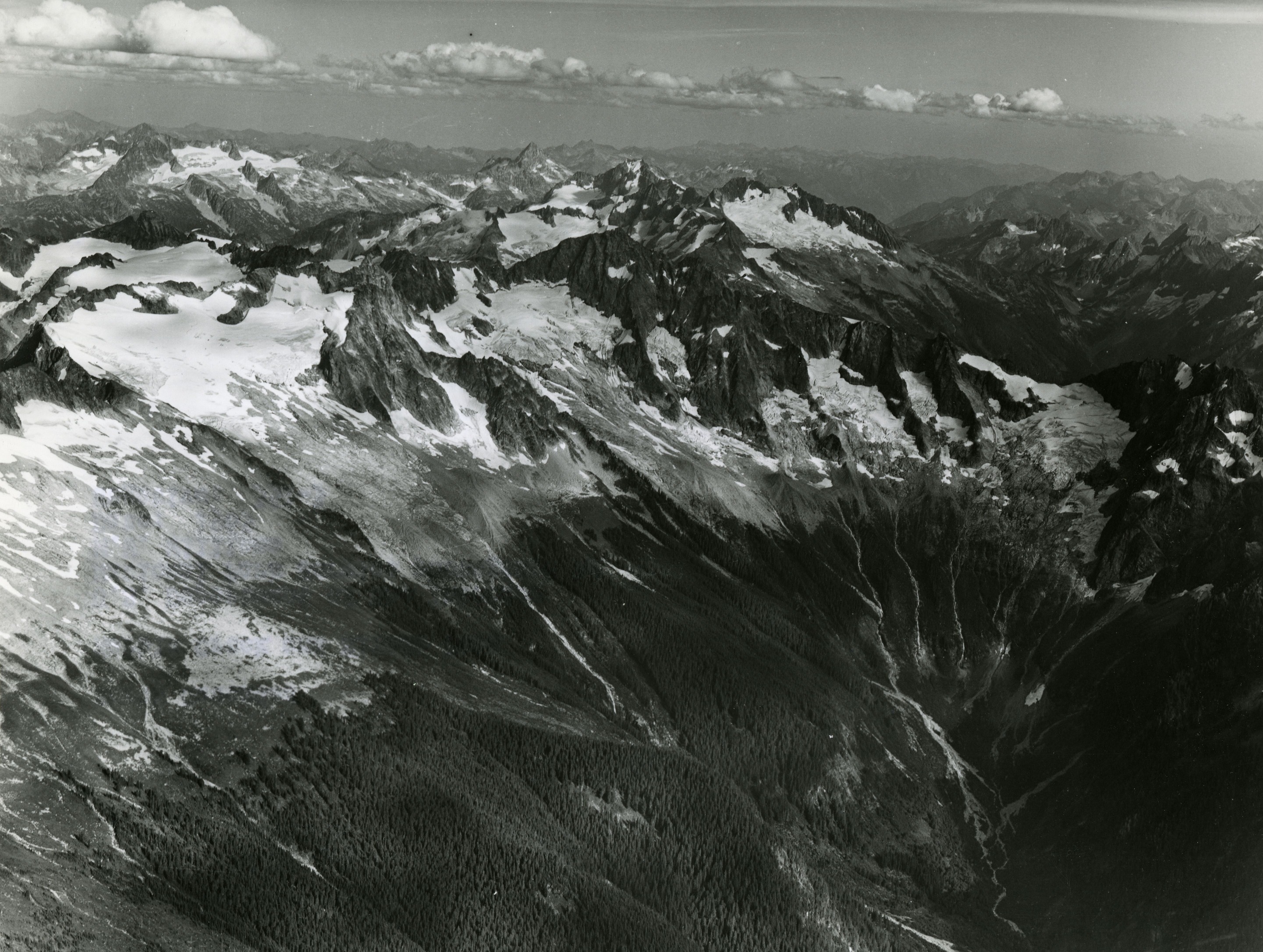 Aerial view of a mountain ridge. The peaks of the mountains are covered in glaciers. The bottom half of the mountains are forested. More mountain peaks can be seen in the background.