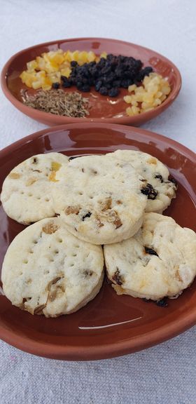 A plate of nice, circular cookies with chunks of dried fruit popping out. Behind it, a plate heaped with seeds and piles of dried, colorful fruits. 