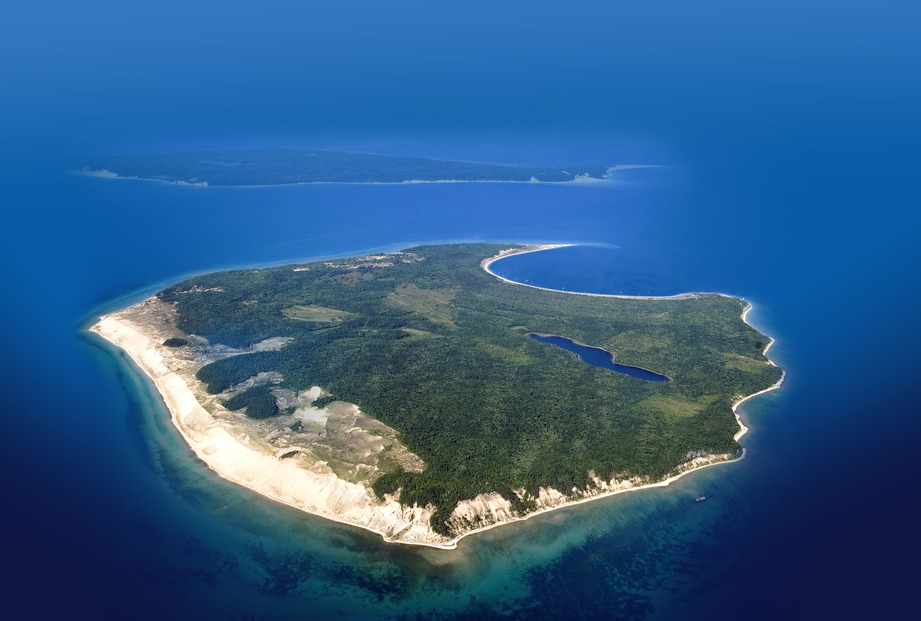 An ariel view of a landform with sand, trees, fields, a lake and a semi-circle water covered harbor.