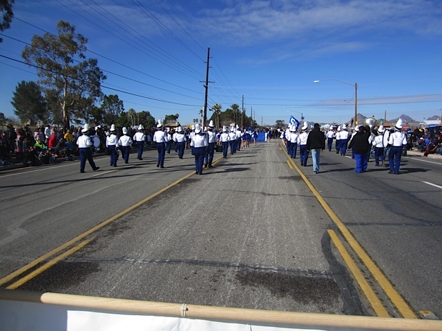 Four rows of people in white jackets and hats and blue pants in a marching band play while walking down a road