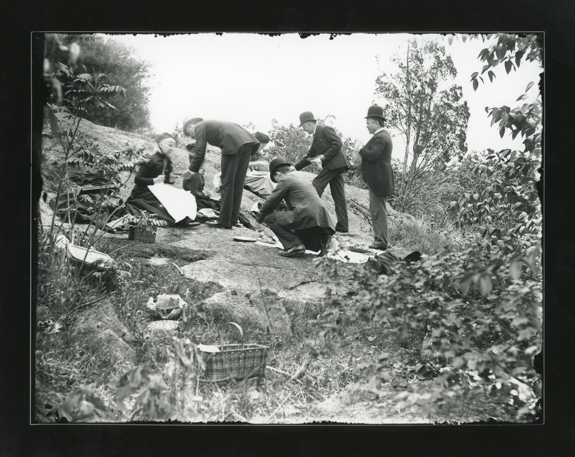 Four white men and four women in full formal dress picnic on a large flat rock in the woods.