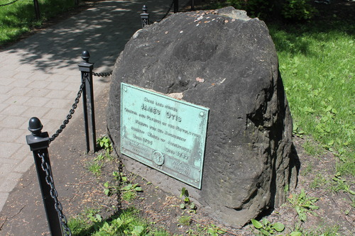 An outdoor image of the grave marker for James Otis. The marker is a large, irregular shaped dark stone. It has a small rectangular plaque on the front of it with text on it including his name and other information that is too small to discern. The ground around the marker is a mix of grass and dirt. There are black fence poles and chains in between around the edge of the walkway that is located  around the marker.