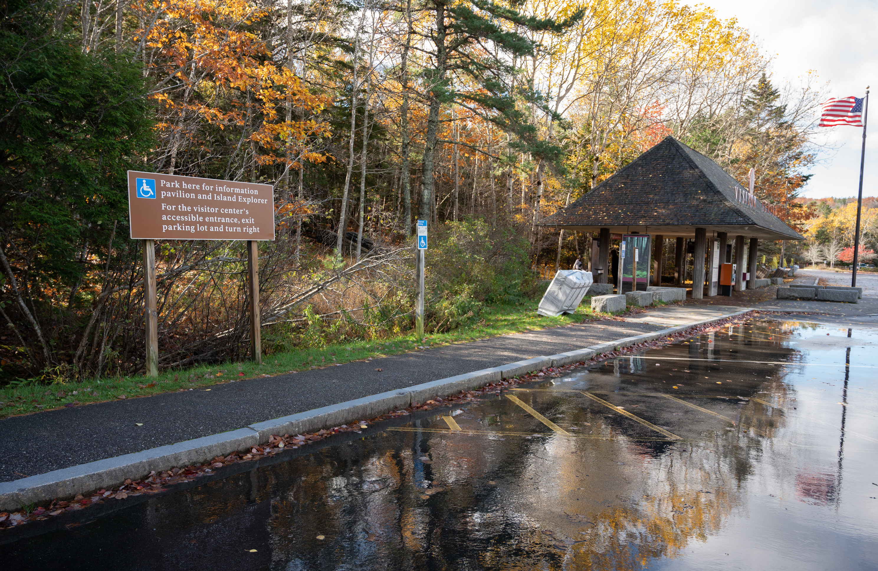 In a forested area, an asphalt parking lot wet from recent rain has a granite curb and asphalt walkway leading to a covered pavilion and flag pole.