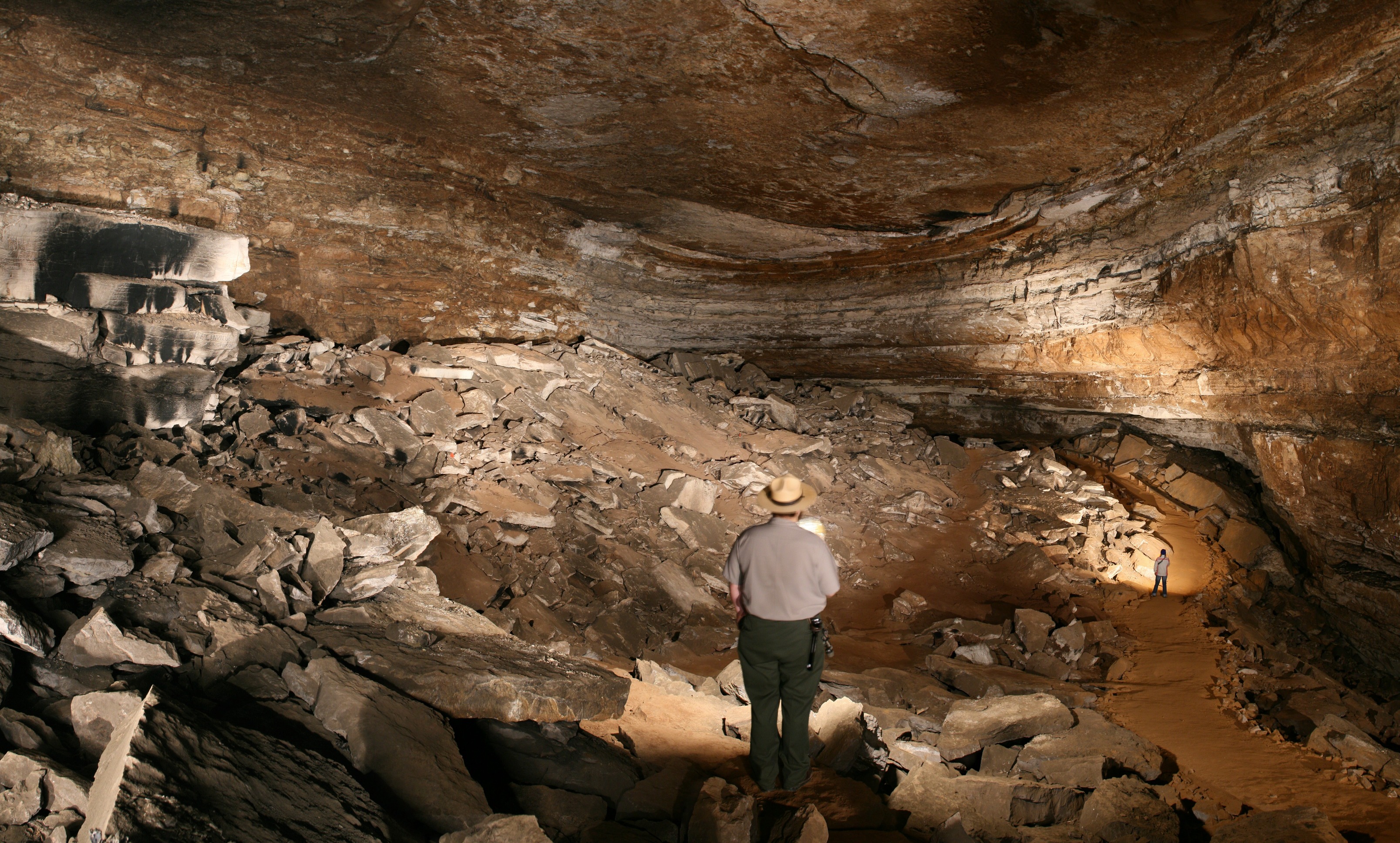 A park ranger stands on a rock lighting up a large cave room. 