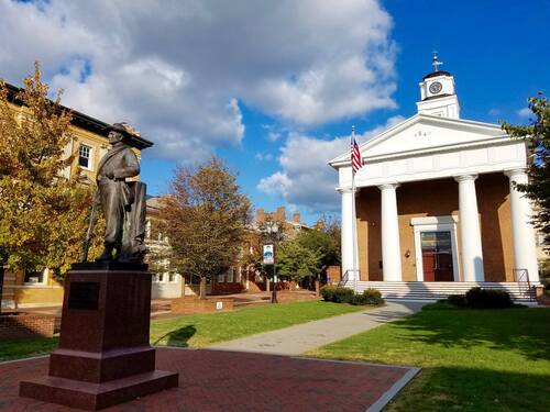 The historic Frederick County Courthouse, a large two-story building with four tall Greek-revival columns in front. A short, paved path leads from the front steps to a stone monument depicting a Confederate soldier holding a rifle at his side. 