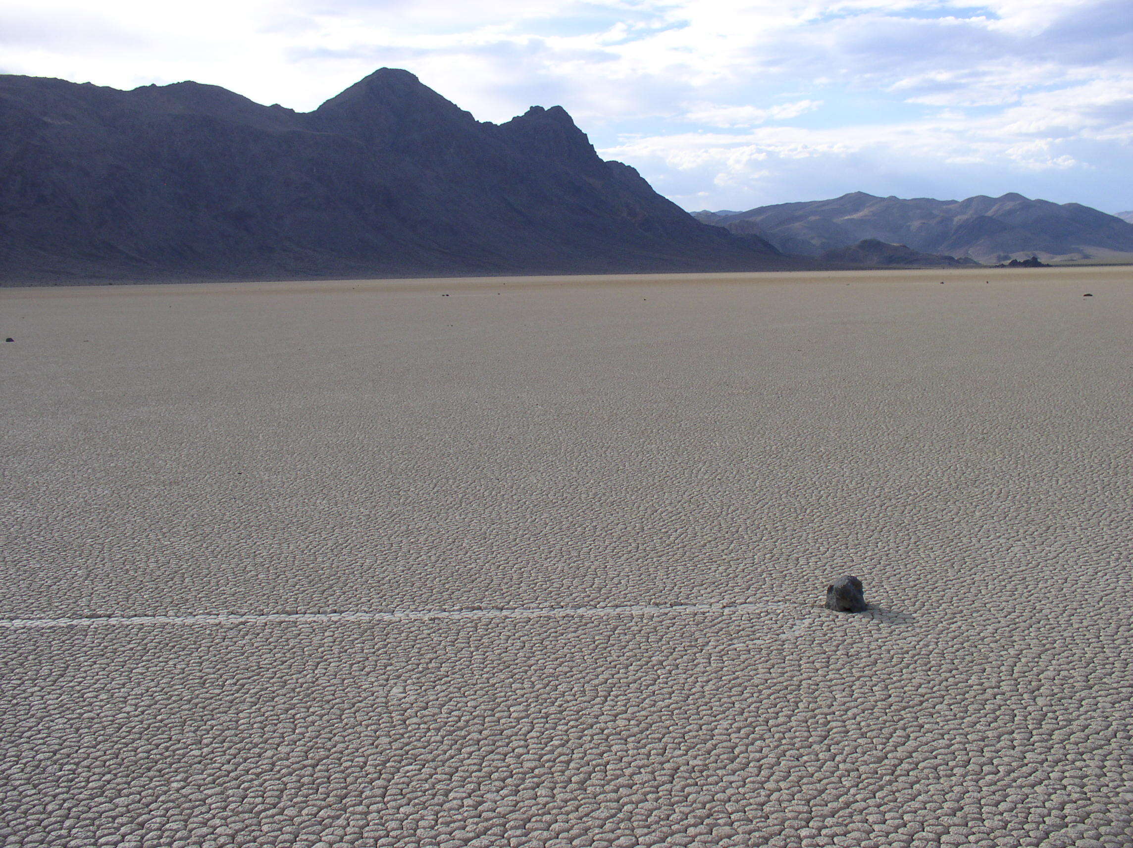 A small rock and a trail of dragged earth behind it on a dry lake bed, with mountains in the distance.