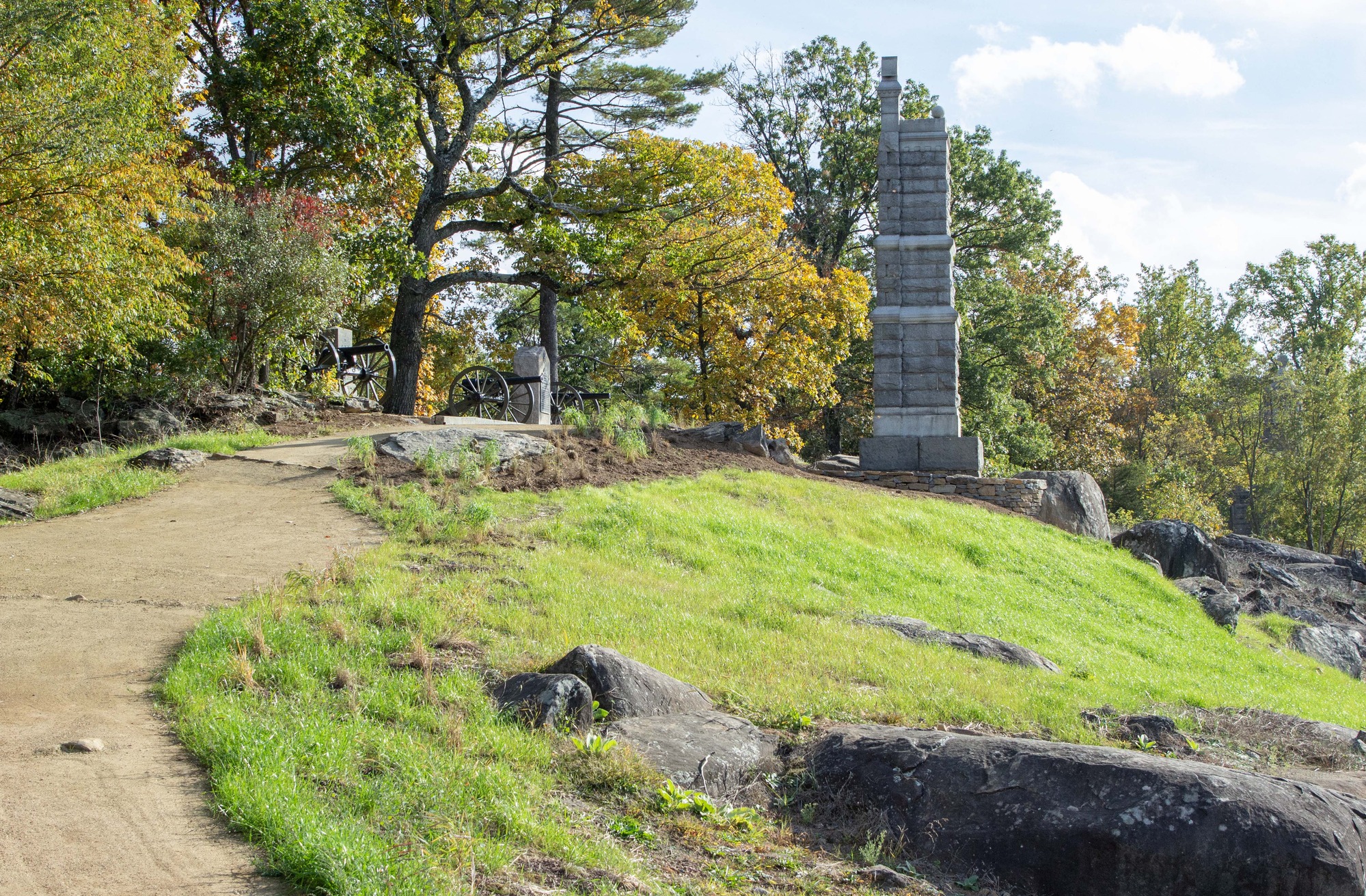 A dirt trail winds past large boulders along the left of the photograph. A grassy knoll lies to the right below a tall grey stone monument.